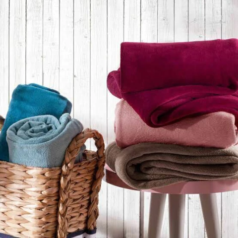 Mantas de microfibra lisa em várias cores dobradas e enroladas em cesta de palha e sobre mesa rosa