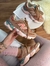 Tênis New Balance 9060 marrom com detalhes em rosa e azul, sendo usado por pessoa com calça rosa clara, sentado próximo a mesa branca com livro
