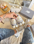 Uma mão segurando um par de tênis New Balance 9060 bege, com um cupcake ao lado, em um ambiente moderno e estiloso.