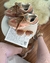 Tênis New Balance 9060 marrom com detalhes em rosa e azul claro, exibido sobre uma mesa redonda branca ao lado de um par de óculos de sol com armação marrom clara