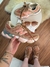 Tênis New Balance 9060 marrom com detalhes em rosa e azul sendo segurado por uma pessoa com tatuagem no braço, ao fundo outro tênis e óculos de sol sobre mesa branca