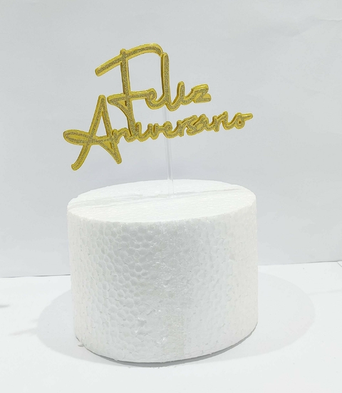 Topo de bolo em formato circular com a inscrição "Feliz Aniversário" em dourado.