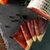 Bresaola maturada artesanal fatiada sobre base de pedra com grãos de pimenta preta e tomate ao fundo