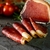 Bresaola maturada artesanal fatiada disposta sobre superfície escura com tomate e ervas ao fundo