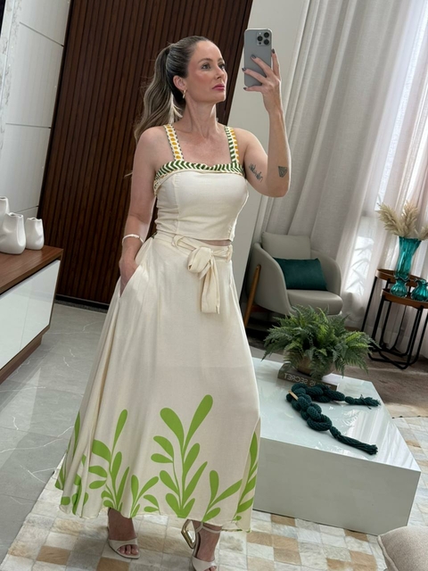 Mulher usando conjunto feminino elegante composto por top branco com detalhes coloridos e saia longa creme com estampa verde de folhas, posando para selfie em ambiente interno moderno.