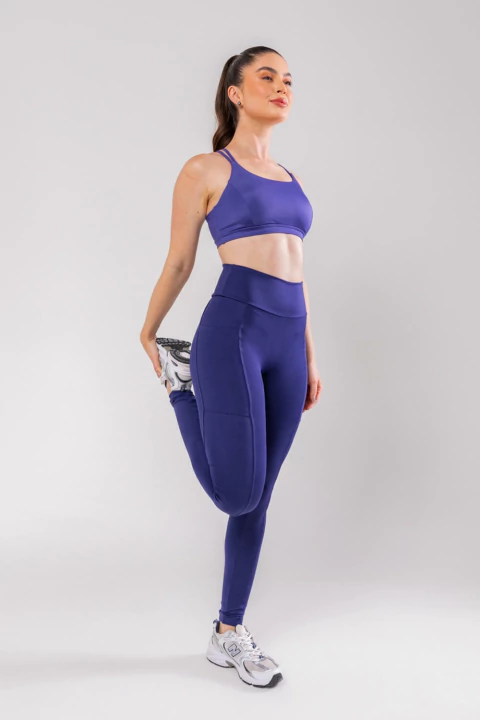 Mulher vestindo calça legging azul escura e top esportivo combinando, realizando alongamento de perna em fundo branco