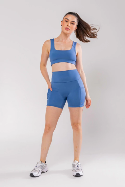 Mulher vestindo bermuda básica fit jeans azul com bolsos, combinada com top esportivo azul e tênis branco