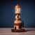 Farol decorativo de madeira com luz acesa, em formato de torre listrada com detalhes em marrom e bege, acompanhado por três árvores pequenas também de madeira
