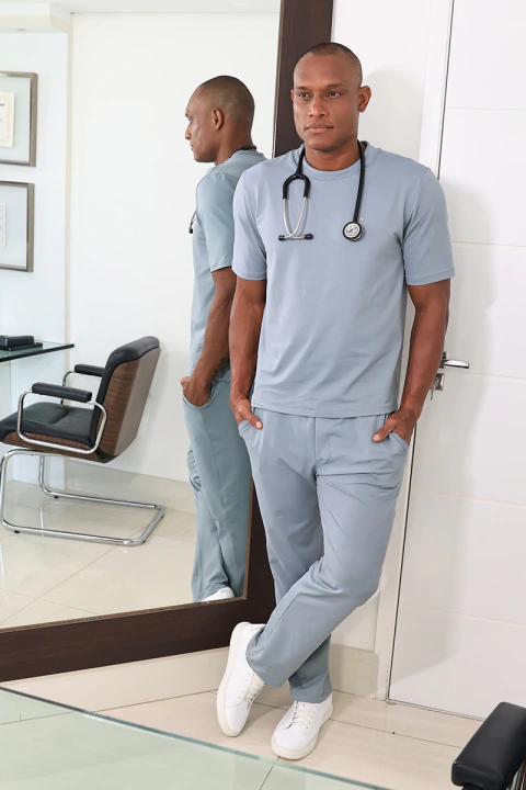 Homem vestindo scrub masculino cinza claro Roma, com estetoscópio no pescoço, encostado em espelho em ambiente clínico moderno