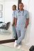 Homem vestindo scrub masculino cinza claro Roma, com estetoscópio no pescoço, encostado em espelho em ambiente clínico moderno