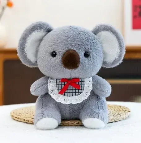 Peluche de koala gris con babero blanco y moño rojo sentado sobre una superficie blanca