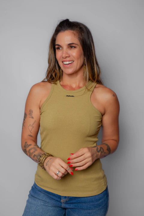 Mulher sorridente vestindo regata nadador canelada bege com jeans azuis, acessórios dourados e unhas vermelhas