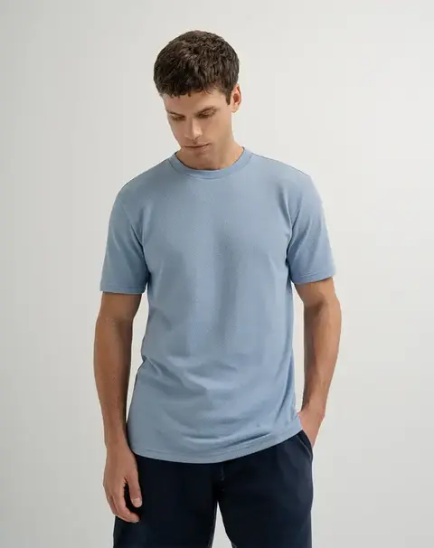 Hombre joven usando camiseta manga corta azul claro y pantalones oscuros, fondo gris claro