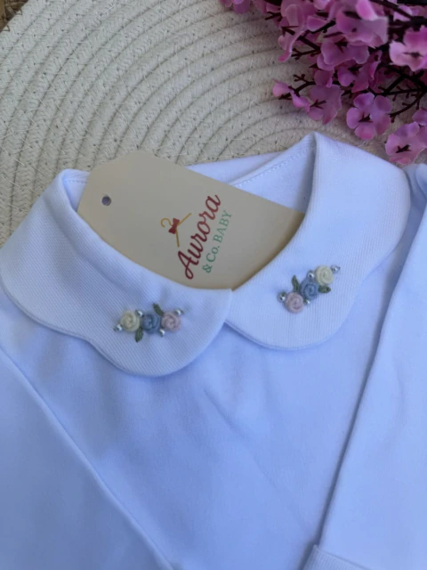 Detalhe da gola arredondada branca de body de bebê com bordados delicados de pequenas flores em tons pastel e pérolas, com etiqueta da marca Aurora & Co. BABY