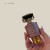 Frasco de perfume feminino pequeno com design de robô em tons dourado e rosa metálico, sendo segurado por uma mão com unhas feitas