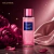 Frasco do body splash Victoria's Secret Pure Seduction Starlit com líquido rosa e partículas brilhantes, ao lado de flores cor-de-rosa e tecido acetinado rosa, em fundo estrelado e ambiente luxuoso