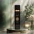 Frasco preto elegante do Body Splash Asad Black Isabelle la Belle em pedestal de mármore verde com fundo desfocado de parede texturizada e decoração com vaso de flores brancas