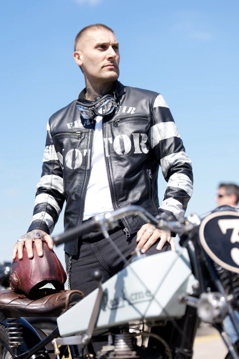 Campera de cuero negra Inmate Racing Jacket, con franjas blancas y letras pintadas a mano, posando junto a una moto de carreras clásica, sosteniendo un casco vintage