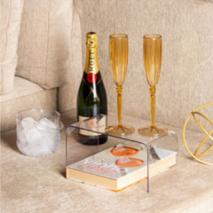 Suporte multiuso diamond 28,5cm x 13,5cm x 13cm no sofá com 2 taças de espumantes em cima e 1 livro em baixo ao lado de uma garrafa de espumante e balde de gelo.
