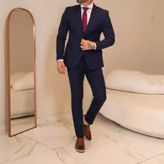 Hombre vestido con traje formal azul marino, camisa blanca y corbata roja, usando zapatos marrones en un ambiente interior con espejo y sofá beige