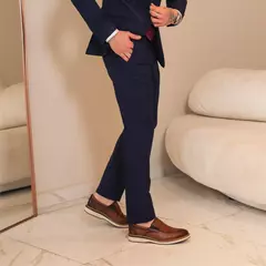Hombre vestido con un traje azul oscuro y zapatos marrones, posando junto a un espejo en un ambiente moderno.