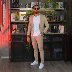 Hombre posando en un ambiente moderno, vistiendo un traje claro y gafas de sol, con una decoración de plantas y objetos en el fondo.