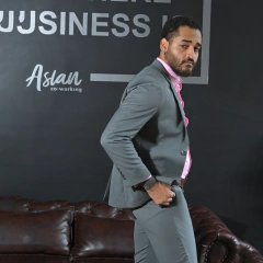 Hombre vestido con traje gris y camisa rosa, posando con una mano en el bolsillo, en un entorno de oficina moderna.