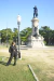 Mulher usando calça pantalona preta e camisa preta, posando ao ar livre próximo a um poste de luz e uma estátua histórica em praça ensolarada