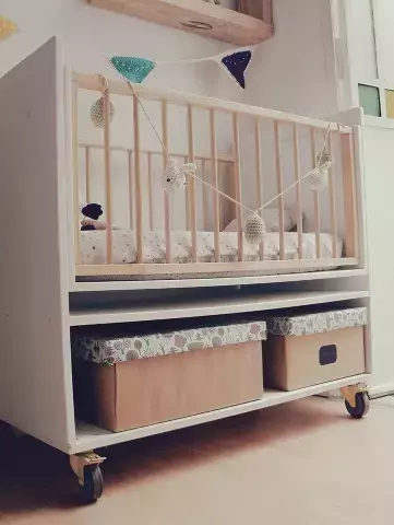 Moisés Colecho Benito, un moisés blanco con barandas de madera, decorado con una guirnalda y almacenamiento inferior.