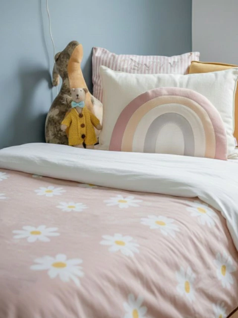 Acolchado infantil rosa con estampado de margaritas blancas sobre cama con almohadones decorativos y peluches