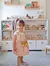 Niña sonriente con un vestido rosa y una cámara de juguete, frente a un mueble de guardado con juguetes y libros.