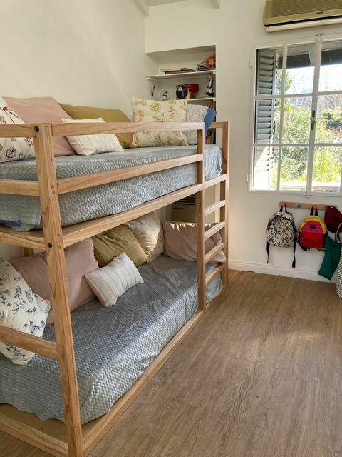 Cama doble tipo litera de madera clara con barandas y escaleras, colchones con cubrecamas celestes y varios almohadones, en habitación luminosa con ventana y piso de madera