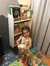 Niña sonriente sosteniendo un libro frente a una biblioteca de pared con varios libros infantiles.