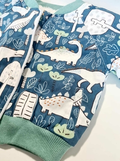 Jaqueta bomber com estampa de dinossauros em fundo azul e detalhes verdes.