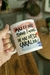 Caneca personalizada branca com a frase "Mas eu não tenho 1 minuto de paz nesse caralho", segurada por uma mão. Fundo desfocado com mesa de madeira e plantas.