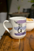 Caneca personalizada branca com alça em formato de coração, decorada com pinceladas em tons de roxo e azul e texto "TEU CU" em preto