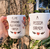 Conjunto de duas canecas brancas com frases divertidas sobre amor, seguradas por uma mão, em um ambiente natural.